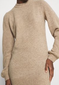 Abito in maglia beige con scollo a girocollo a coste e maniche lunghe leggermente a sbuffo. Tessuto morbido e forma aderente.
