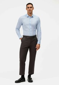 Chemise bleu clair à boutons avec un motif subtil, associée à un pantalon sur mesure brun foncé et des chaussures noires, créant un look décontracté chic.