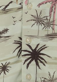 Tela verde claro con un estampado tropical de palmeras marrones y rosas, aves, y botones color crema en el centro.