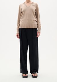 Beige stickad tröja med V-ringning, ribbat nederkant och muddar, i kombination med löst sittande svarta byxor och platta svarta sandaler.