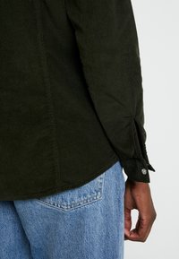 Mörkgrön kordflorse skjorta med texturerad tyg, snedställd ärmdetalj och silverknapp, i kombination med ljusblå denimjeans.