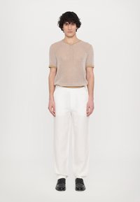 Pull en tricot beige à manches courtes et finitions claires, associé à un pantalon blanc ample et des chaussures noires. Fond uni.