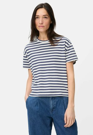 Vrouw met lang donker haar, gekleed in een blauw-wit gestreept T-shirt met korte mouwen en losse blauwe jeans, staand met één hand in haar zak.
