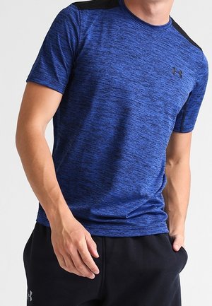 Mann trägt ein blaues, strukturiertes, kurzärmeliges Sportshirt und schwarze Jogginghosen, steht mit einer Hand in der Tasche.