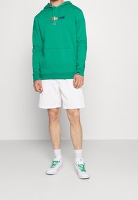 Felpa verde realizzata in un tessuto morbido, con un logo multicolore. Abbinata a pantaloni corti bianchi e sneakers bianche con dettagli verdi.