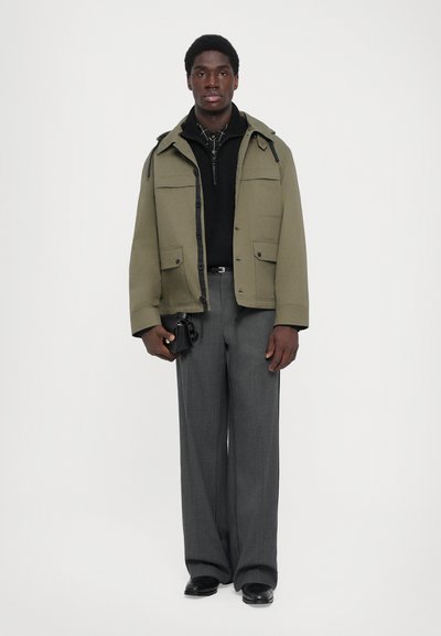Olivgrüne Jacke mit vier Fronttaschen, schwarzer Rollkragenpullover, graue Hose und schwarze Schuhe. Hält eine kleine schwarze Tasche.
