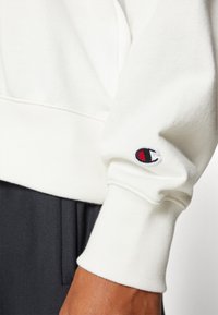 Felpa bianca con polsini a coste; presenta un piccolo logo ricamato in rosso e blu sulla manica; texture morbida visibile.