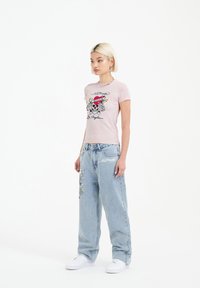 Růžové tričko s krátkým rukávem a grafikou lebky a textem; spárováno se světle modrými širokými džínami se vyšívanými detaily; bílé tenisky.