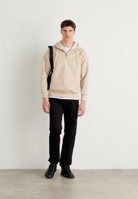 Haut zippé crème avec cordons de serrage, pantalon noir et chaussures noires. Le modèle se tient avec un sac à dos. Texture lisse, design minimaliste.