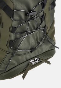 Sac à dos vert avec un tissu texturé, un design de cordon élastique réglable et un matériel de fermeture éclair métallique avec des languettes à la base.