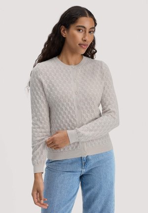 Frau mit langen dunkel lockigen Haaren, die eine hellgraue gemusterte, geknöpfte Strickjacke und hellblaue Jeans trägt, und den Ärmel am linken Arm hochkrempelt.