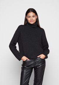 Schwarzer, gerippter Rollkragenpullover mit lockerer Passform, langen Ärmeln und verkürztem Saum, kombiniert mit glänzenden schwarzen Kunstlederhosen.