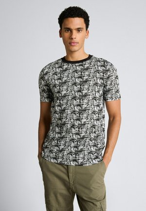 Jonge man met een zwart-wit T-shirt met palmboommotief en olijfgroene broek, staand met handen in de zakken tegen een effen achtergrond.