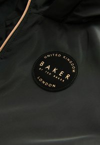 Svart jacktyg med en cirkulär svart patch med guldtext. Patchen har texten "BAKER by TED BAKER, LONDON" och inkluderar platsinformation.