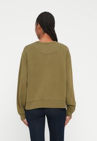 Felpa verde oliva con taglio rilassato, caratterizzata da un collo rotondo, cuciture delle spalle scese e polsini a costine. Materiale morbido e texturizzato.