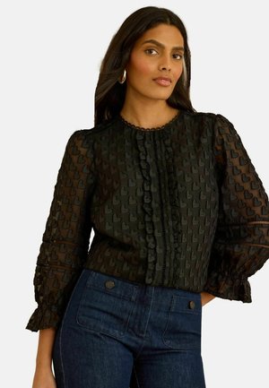 Kvinde iført en gennemsigtig sort bluse med hjerteprint, flæset front og manchetter, kombineret med mørkeblå jeans med høj talje.