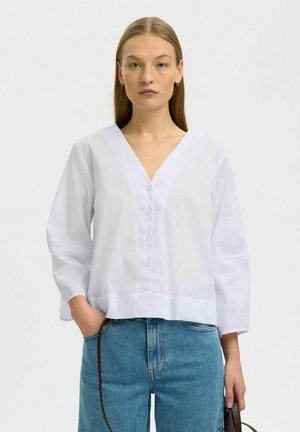 Femme aux cheveux blonds lisses portant une chemise blanche à col en V boutonnée et un jean bleu, tenant un sac à main à sangle tressée marron.