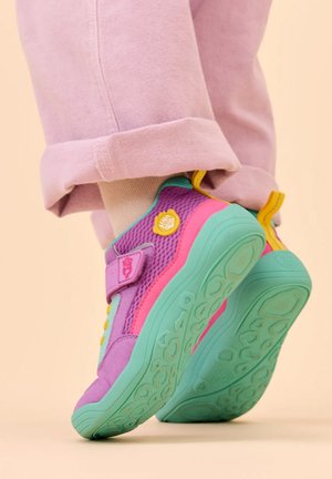 Kind draagt kleurrijke paarse, roze en blauwgroene sneakers met gele accenten en beige sokken, staand op tenen tegen een effen achtergrond.