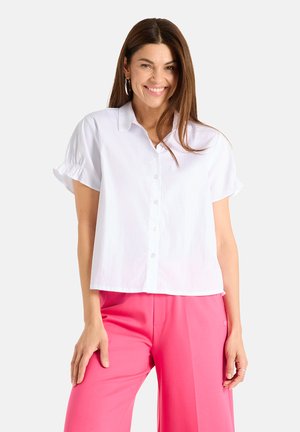 Woman smiling wearing a short-sleeve white button-up shirt and bright pink pants, standing against a plain light background.