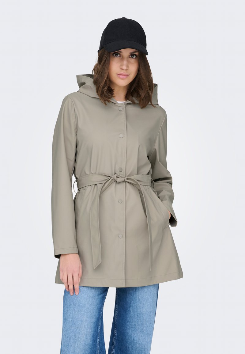 Mujer con impermeable beige con cinturón y capucha, gorra de béisbol negra y vaqueros azules, de pie con una mano en el bolsillo.