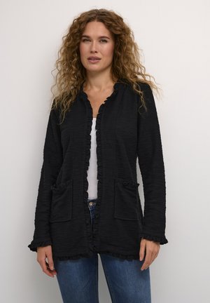 Schwarze Strickjacke mit Rüschensaum, langen Ärmeln, Taschen, strukturiertem Stoff und offenem Design, getragen über einem weißen Oberteil und blauen Jeans.
