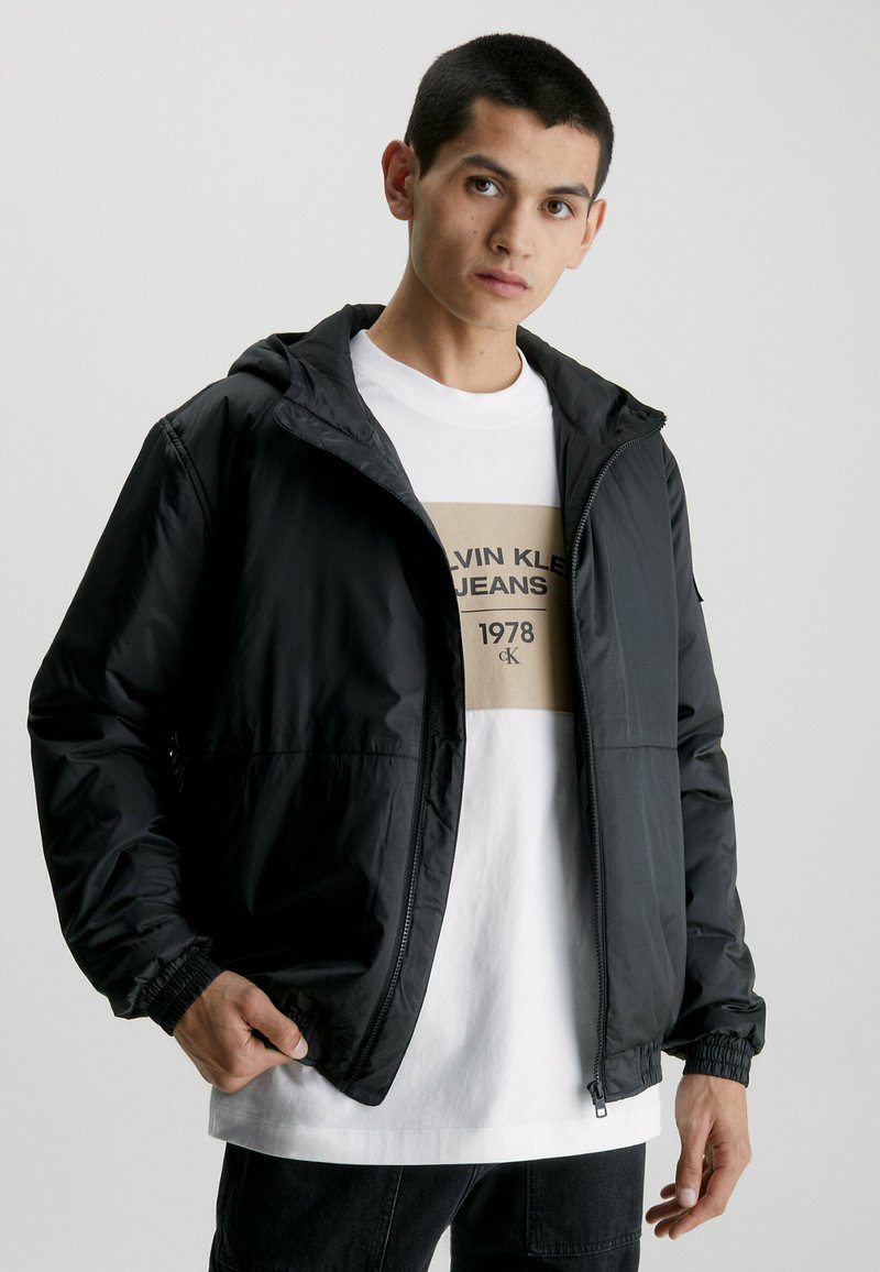 Giacca nera in nylon con cappuccio, chiusura a zip, polsini elasticizzati e tasche laterali, indossata sopra una maglietta bianca con una grafica beige.