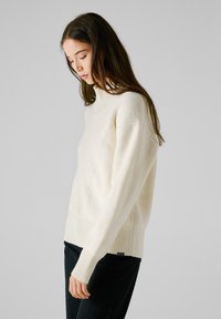 Elfenbensfärgad stickad tröja med hög krage, ribbade ärmslut och nederkant, med diagonala texturerade mönster och en lös passform över svarta jeans.