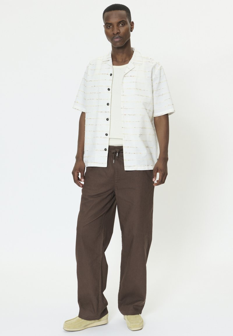 Chemise blanche à manches courtes avec des boutons noirs et de fines lignes horizontales, associée à un pantalon marron ample et des chaussures plates crème.