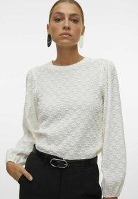 Blusa bianca texturizzata con maniche lunghe e spalle a sbuffo, abbinata a una cintura nera con fibbia argentata, indossata su pantaloni neri.