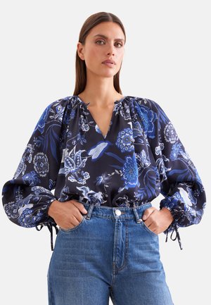 Femme portant un chemisier bleu marine à motifs floraux avec des manches longues bouffantes et un jean bleu, debout les mains dans les poches devant un fond uni.