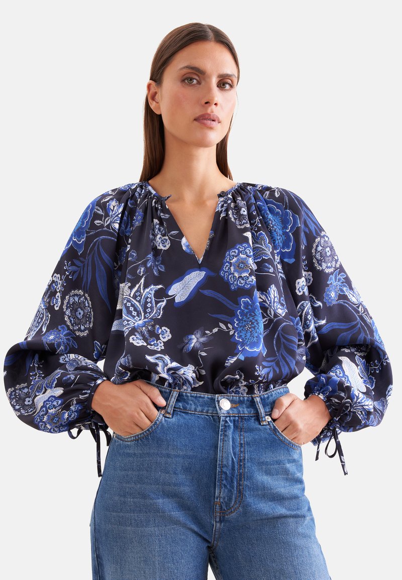 Femme portant un chemisier bleu marine à motifs floraux avec des manches longues bouffantes et un jean bleu, debout les mains dans les poches devant un fond uni.