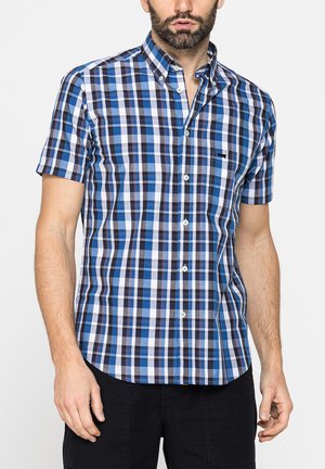 Uomo che indossa una camicia a quadri blu, nera e bianca a maniche corte con pantaloni neri su uno sfondo semplice.