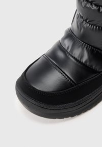 Gros plan sur une botte d'hiver noire isolée avec une tige rembourrée et cousue et une semelle en caoutchouc texturé sur un fond blanc.