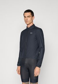 Black cycling jacket in lightweight fabric with a high collar, front zipper, and logo on the chest. Worn over fitted cycling shorts.