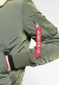 Tmavě zelená bomber bunda z hladkého nylonu s ribovanými manžetami. Obsahuje zipovou kapsu na rukávu a červenou štítku "ODSTRAŇTE PŘED LETEM".