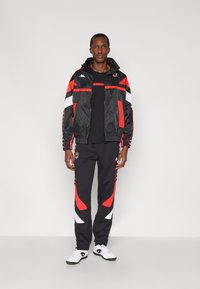 Czarna i czerwona kurtka sportowa z białymi akcentami, zestawiona z czarnymi spodniami z czerwono-białymi wzorami oraz białymi butami sportowymi.