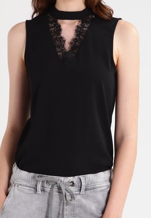 Czarny top bez rękawów z dekoltem w serek wykończonym koronką i kołnierzem choker, zestawiony z jasnoszarymi spodniami ze sznurkiem.