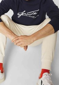 Personne assise, les mains jointes, portant un sweat-shirt bleu marine avec un logo blanc, un pantalon beige, des chaussettes rouges et des baskets blanches avec des accents noirs.