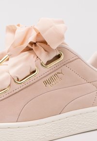Les baskets Puma Basket en daim rose clair présentent une surface texturée, des œillets dorés et un grand nœud en satin pour les lacets. Semelle en caoutchouc blanche.