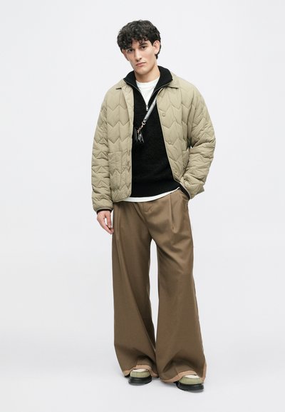 Κουβερτίνα ελαιοπράσινη ζακέτα, μαύρος υφασμάτινος πουλόβερ, φαρδιά μπεζ παντελόνια, μαύρα παπούτσια με πράσινες λεπτομέρειες, όρθιος σε λευκό φόντο.