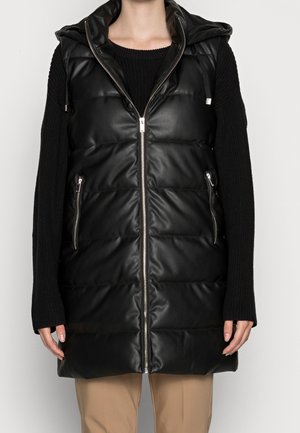 Personne portant un gilet matelassé sans manches noir à capuche avec une fermeture éclair avant et des poches zippées sur un pull noir à manches longues et un pantalon beige.
