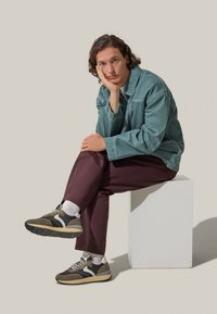 Giacca di denim teal, camicia bianca, pantaloni bordeaux e sneakers multicolori con accenti grigi e verdi. Pedestallo geometrico semplice.