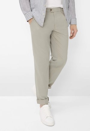 Uomo con pantaloni beige, sneaker bianche, camicia bianca e camicia grigia a righe con bottoni, in piedi davanti a uno sfondo bianco.