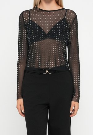 Mujer con top negro transparente de manga larga con lunares blancos sobre un sostén negro, combinado con pantalones negros y cinturón con hebilla dorada.