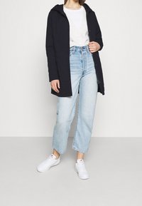 Cappotto blu navy con cappuccio, tessuto leggero, chiuso con bottoni, abbinato a un cardigan bianco, jeans a vita alta azzurri chiari e sneakers bianche.