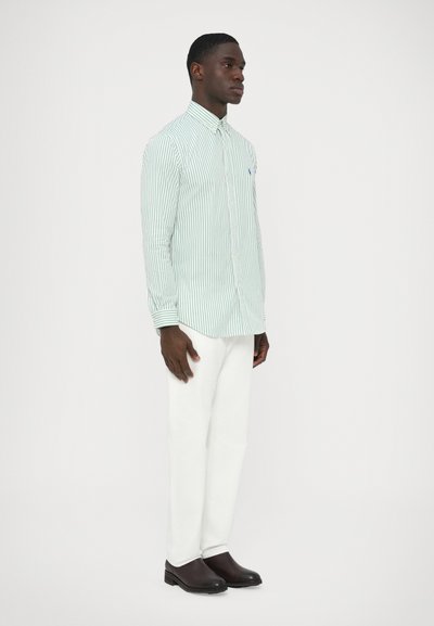 Zeleno-bela črtasta srajca s srajčnim ovratnikom, dolgimi rokavi in majhnim logotipom, kombinirana z belimi hlačami in temnimi čevlji.