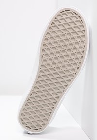 Podeszwa białego sneakersa z beżowym, waflowym wzorem na tle białym.