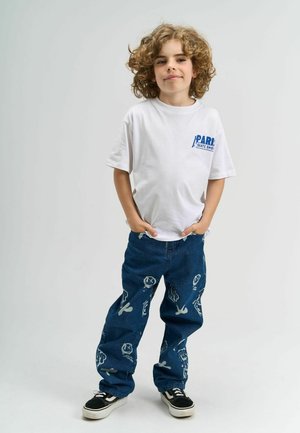 Tricou alb cu text albastru "PARIS SKATE SHOP", asortat cu blugi din denim albastru ce afișează desene grafice albe și adidași negri.