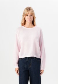 Young woman with blonde hair wearing a light pink knit sweater and dark blue jeans, standing against a plain white background.
