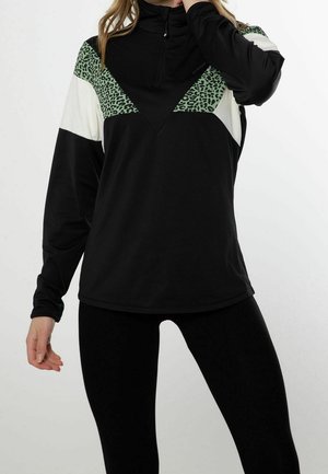 Femme portant des leggings noirs et un pull noir avec un imprimé léopard vert et des panneaux blanc cassé en blocks de couleur, posant avec une main près du cou.